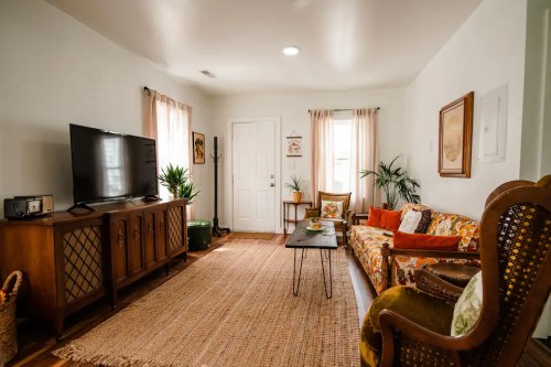 Cozy living room with a patterned sofa, TV, plants, and natural light from windows, featuring a warm, inviting atmosphere.
