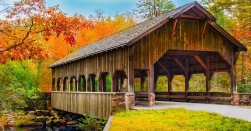 A rustic covered bridge spans a calm stream, surrounded by vibrant trees and greenery under a blue sky.