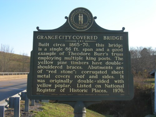 A historic covered bridge sign detailing its history, surrounded by trees and a rural landscape.