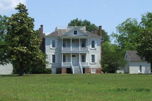 A historic white house with a porch, surrounded by green grass and trees, under a clear blue sky.