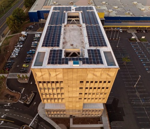 Aerial view of a modern building with solar panels on the roof, surrounded by a parking lot and road.