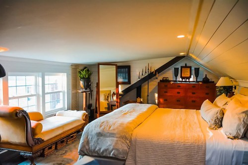 Cozy attic bedroom with a large bed, vintage furniture, and soft lighting, featuring a chaise lounge and decorative accents.