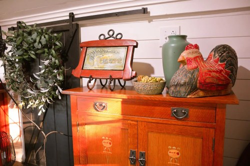 A wooden cabinet with a "Welcome" sign, a green vase, a decorative bowl, and a rooster figurine, surrounded by greenery.