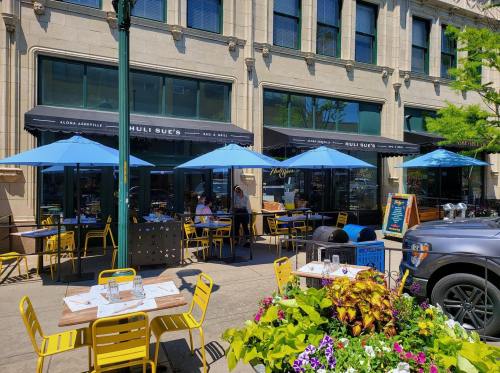 Outdoor dining area of a restaurant with blue umbrellas, yellow chairs, and colorful flowers in front.