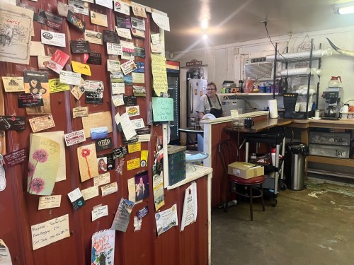 A cozy café interior with a wall covered in notes and flyers, and a smiling barista behind the counter.