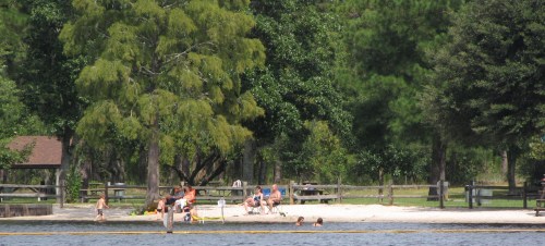 A sandy beach by a lake with people relaxing, swimming, and enjoying the outdoors under trees.
