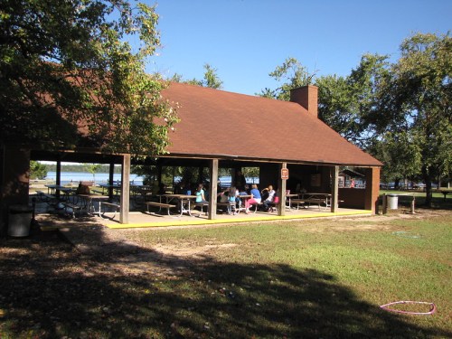 A picnic shelter with a brown roof, surrounded by trees, where people are gathered at tables near a lake.