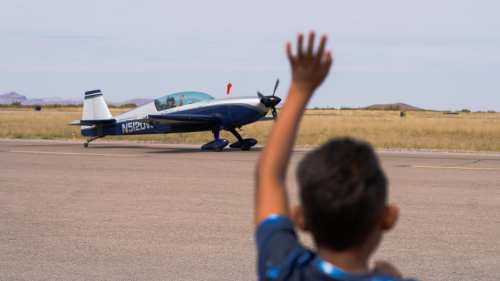 A child waves as a small airplane taxis on a runway in a grassy field.