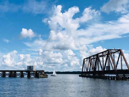 A sailboat glides past a rusty bridge and a wooden pier under a bright blue sky with fluffy white clouds.