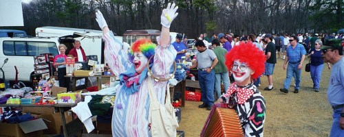 Two clowns in colorful costumes smile and wave at a bustling outdoor market filled with people and various items for sale.
