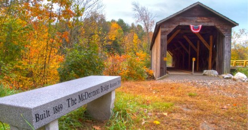 A gray stone bench sits next to a wooden covered bridge with red-white-and-blue bunting that's surrounded by vibrant fall foliage.
