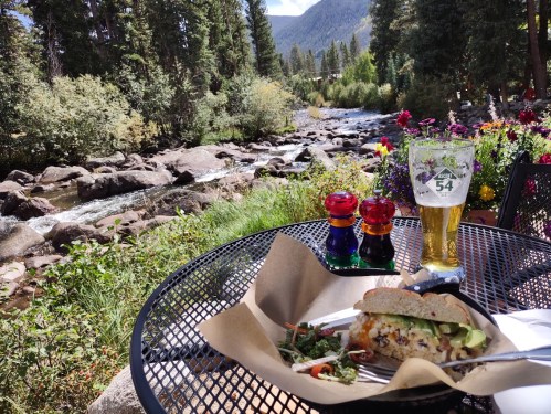 A scenic outdoor dining setup with a sandwich, salad, beer, and a view of a river and mountains.