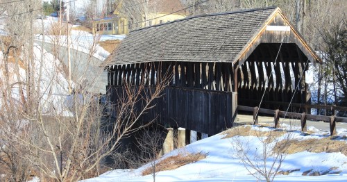 A snowy landscape featuring a covered bridge above a flowing stream, surrounded by rocky terrain and trees with a yellow house in the background.