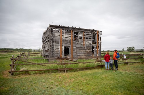 Two people stand near a weathered, abandoned wooden structure surrounded by tall grass and a wooden fence.
