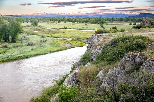 A person stands on a rocky ledge overlooking a river and lush green landscape under a colorful sunset sky.