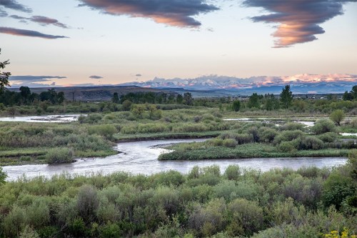 A winding river flows through lush greenery, with mountains and colorful clouds in the background at sunset.