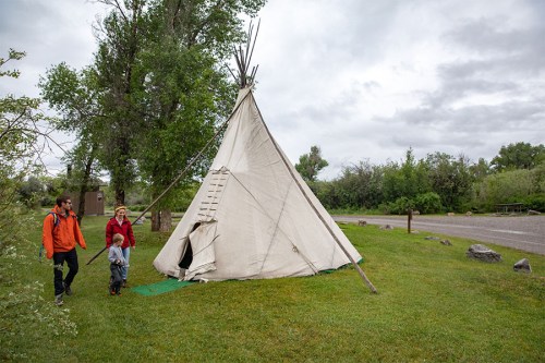 A couple walks past a traditional teepee on a grassy area, surrounded by trees under a cloudy sky.