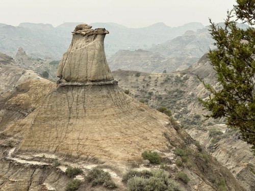 A unique rock formation rises above a rugged landscape, surrounded by hills and sparse vegetation under a hazy sky.
