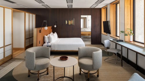 Modern hotel room featuring a bed, two chairs, a round table, and large windows with natural light.