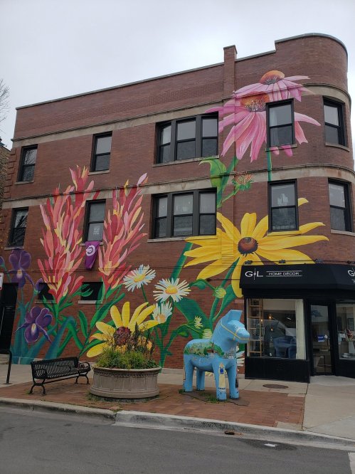 Colorful flower mural on a brick building, with a blue horse sculpture and a bench in front.