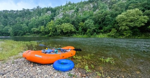 A bright orange raft and a blue tube, sitting on the Ouachita River in Arkansas, surrounded by greenery.