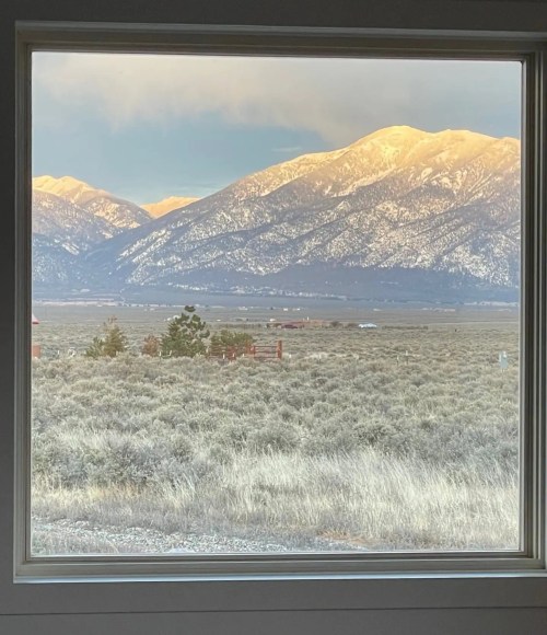 A scenic view of snow-capped mountains framed by a window, with a foreground of sagebrush and open landscape.