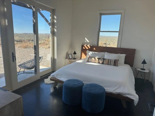 Bright bedroom with a large bed, two blue stools, and a view of the outdoors through glass doors.