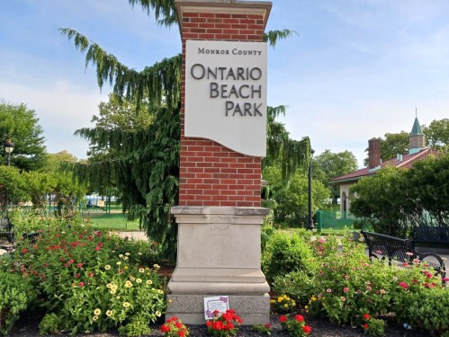 Sign for Ontario Beach Park in Monroe County, surrounded by colorful flowers and greenery on a sunny day.