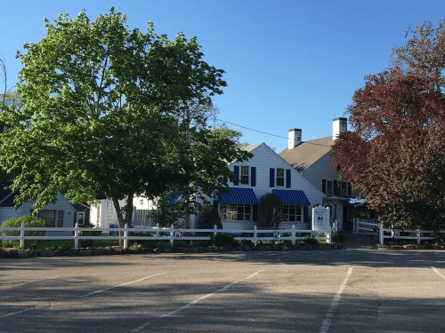 A charming white building with blue awnings, surrounded by trees, in a sunny outdoor setting with a parking lot.