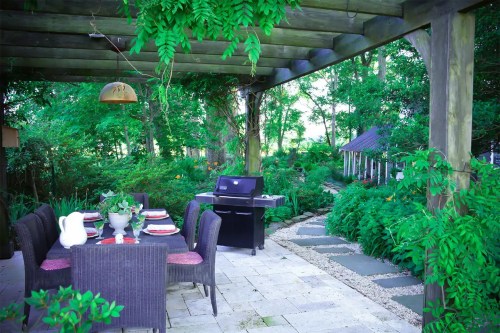 A serene outdoor dining area with a grill, surrounded by lush greenery and a stone pathway.