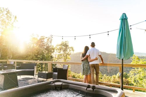 A couple stands together on a deck, overlooking a scenic landscape at sunset, with a hot tub nearby.