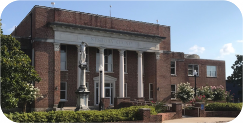 Historic brick building with columns, a statue in front, and trees, representing Neshoba County. Clear blue sky above.