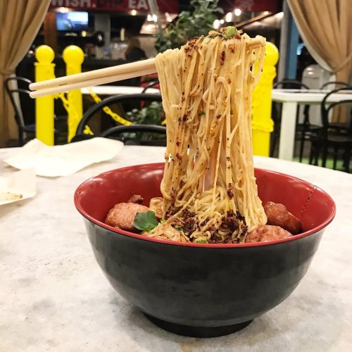 A bowl of noodles topped with meatballs and herbs, with chopsticks lifting a portion of the noodles.