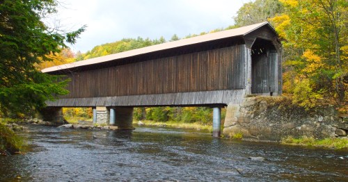 A long weathered, covered bridge crosses a river at an angle with trees starting to turn fall colors in the background.