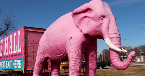 a massive pink elephant made of fiberglass stands in front of a trailer used as a sign for the pink elephant antique mall on route 66 in illinois