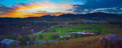 A vibrant sunset over rolling hills and a green valley, with scattered trees and buildings in the foreground.