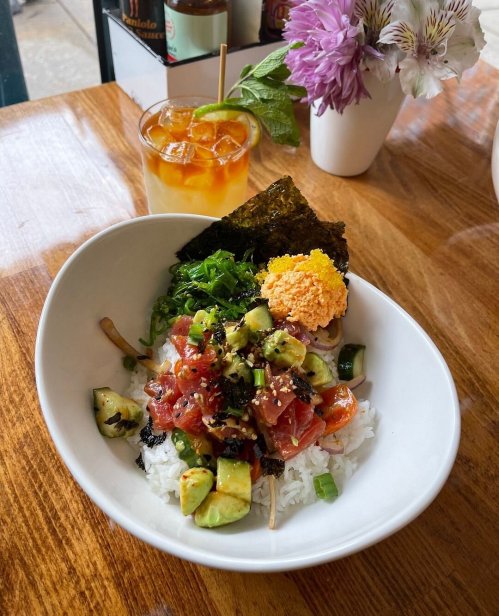 A bowl of rice topped with diced tuna, avocado, seaweed, and orange roe, alongside a refreshing drink and flowers.
