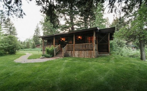 A rustic cabin surrounded by lush greenery and trees, featuring a wooden porch and steps leading to the entrance.