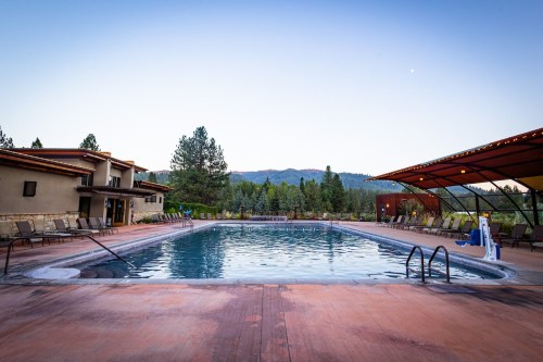 A serene outdoor pool surrounded by trees and mountains, with lounge chairs and a clear sky at dusk.