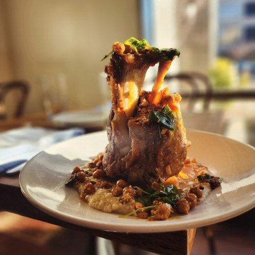 A beautifully plated dish featuring a large lamb shank on a bed of lentils and greens, garnished with herbs.