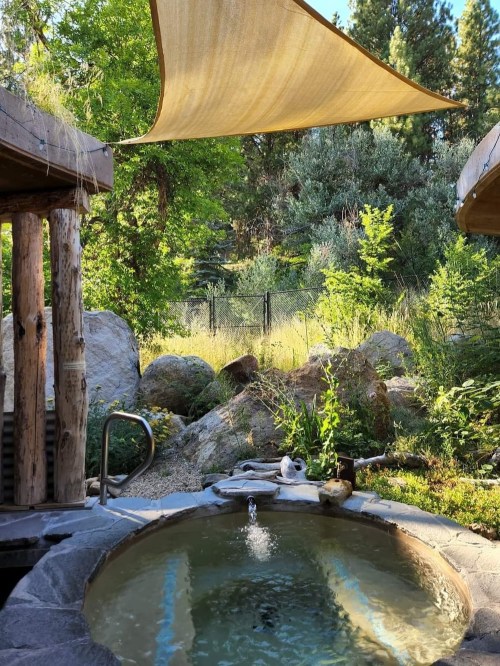 A serene outdoor hot tub surrounded by lush greenery and rocks, with a shade sail overhead.