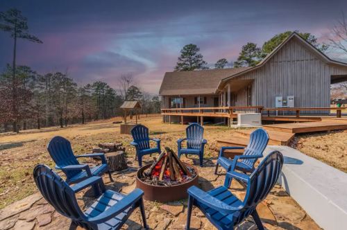 A cozy outdoor fire pit surrounded by blue chairs, with a wooden cabin and trees in the background under a colorful sky.