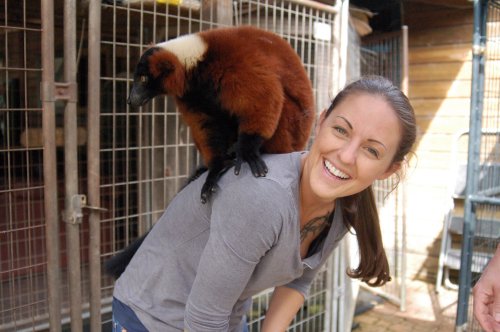 A woman smiles while a brown and white lemur sits playfully on her shoulder.