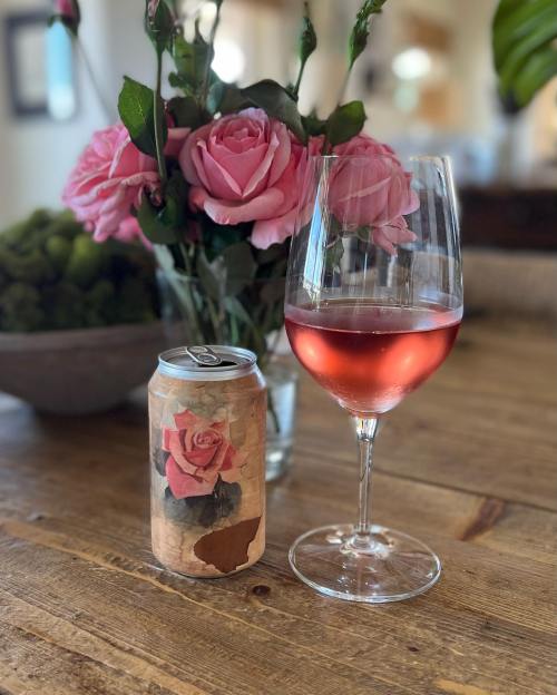 A glass of rosé wine next to a can with a rose design, accompanied by a bouquet of pink roses on a wooden table.