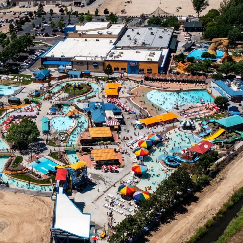 Aerial view of a vibrant water park with pools, slides, and colorful umbrellas, surrounded by trees and parking areas.