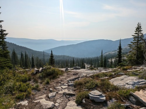 A rocky trail leads through a forested mountain landscape, with hazy hills in the distance under a clear sky.
