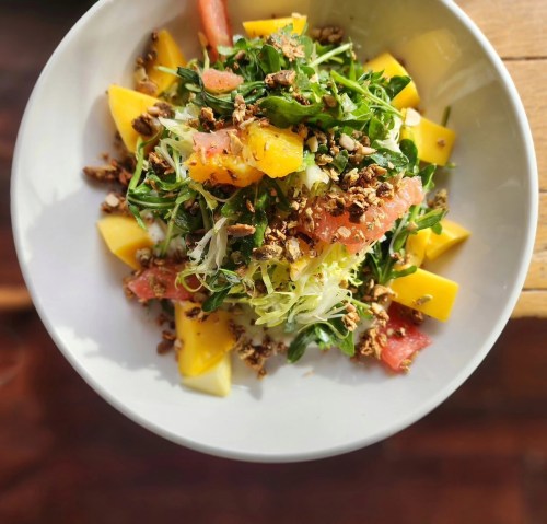 A vibrant salad with mixed greens, mango, grapefruit, and crunchy granola, served in a white bowl.