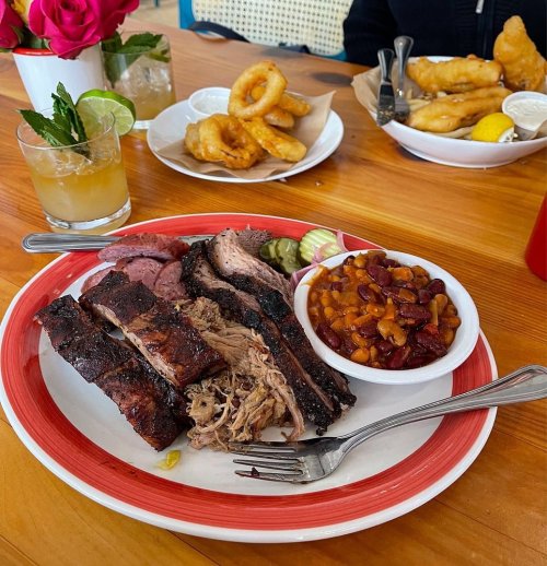 A plate of barbecue featuring ribs, pulled pork, pickles, and baked beans, with drinks and onion rings in the background.