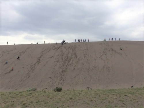 A sandy hill with people climbing and standing at the top, under a cloudy sky. Green grass is visible at the base.