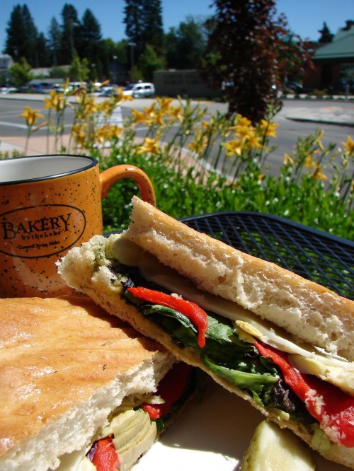 A close-up of a sandwich with vegetables, alongside a coffee mug, set against a sunny outdoor scene with flowers.